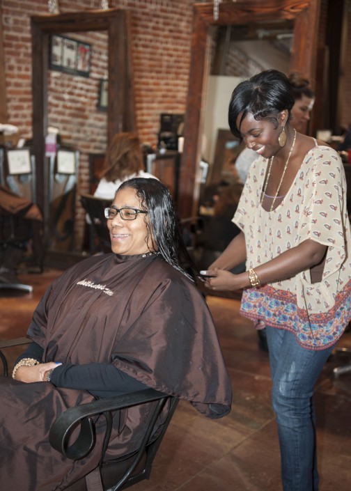 a woman in jeans and a flowy top cutting hair for a woman seated and wearing a brown smock