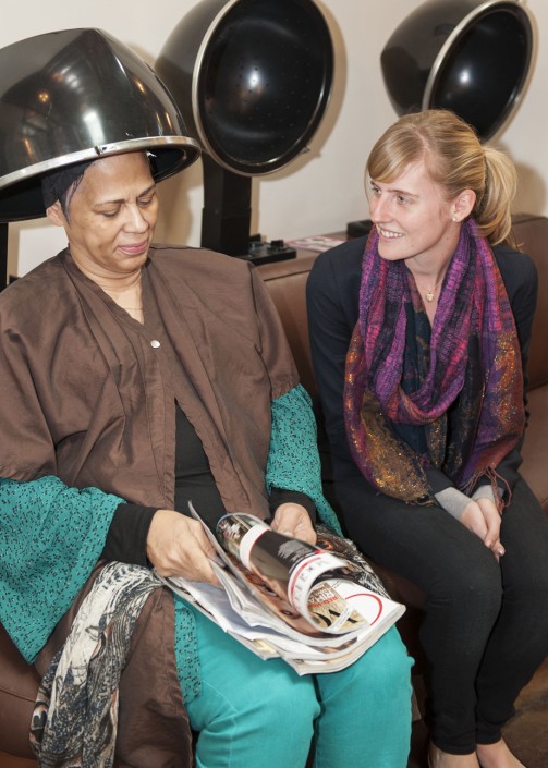 a woman seated under a hair dryer while looking at magazines with another woman seated beside her
