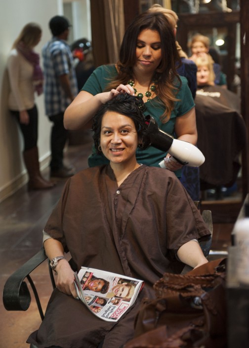 a woman seated while wearing a brown smock holding a magazine while another blow dries her hair