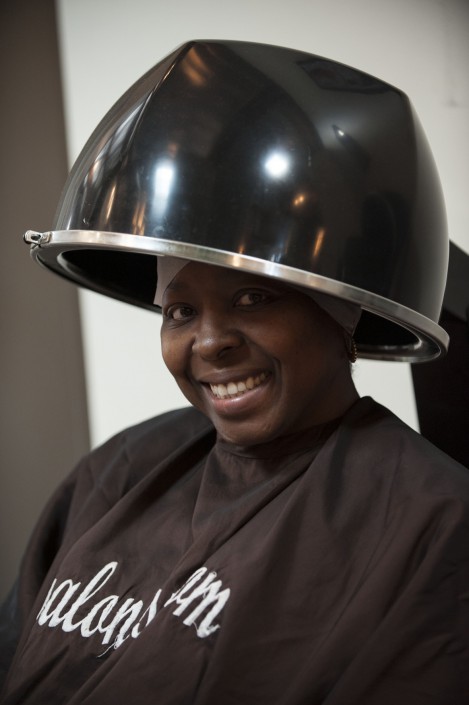 a woman smiling while wearing a brown smock and seated beneath a hair dryer