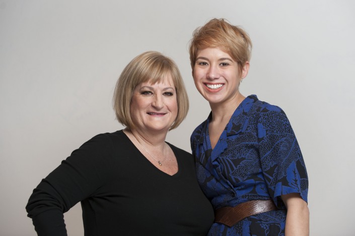 a woman wearing a blue shirt standing beside a shorter woman in a black sweater against a white background