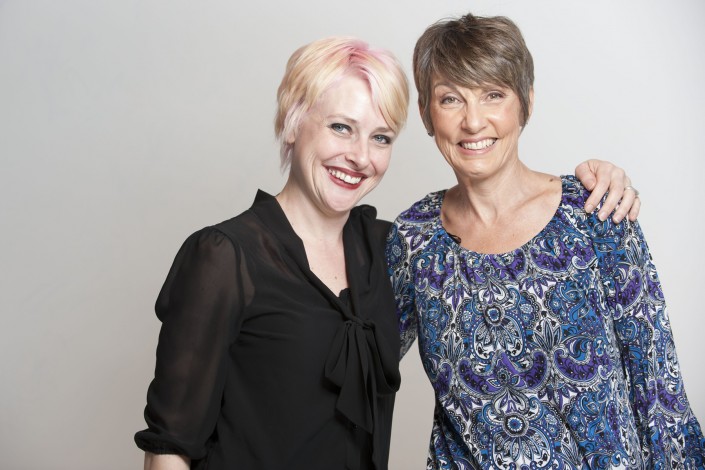 a woman in a black shirt with her arm around a woman in a blue shirt standing against a white background