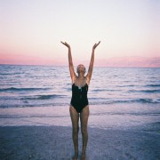 a woman in black bathing suit standing on a beach with her hands in the air