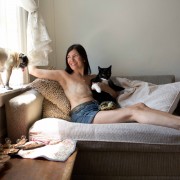 a woman lying on a couch topless wearing jean shorts showing breasts with surgery scars holding a black and white cat with a pug dog on a windowsill