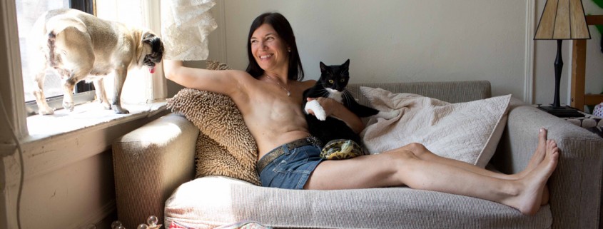 a woman lying on a couch topless wearing jean shorts showing breasts with surgery scars holding a black and white cat with a pug dog on a windowsill