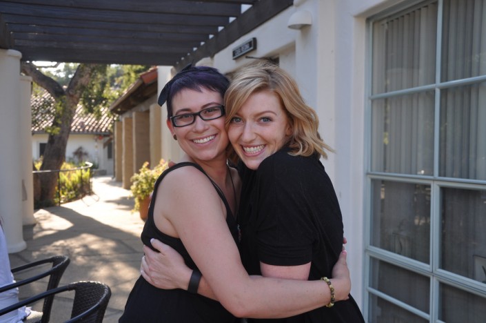 two women wearing black hugging while standing on a patio in front of windows
