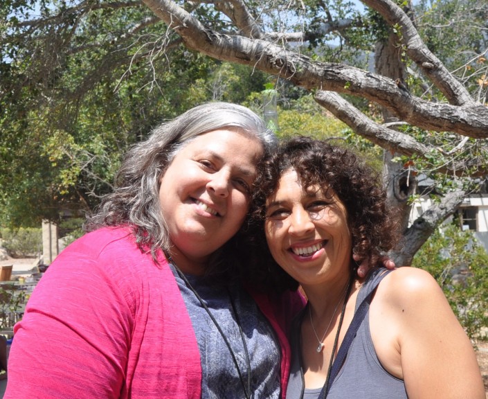 two women standing with their heads together and smiling in front of large trees
