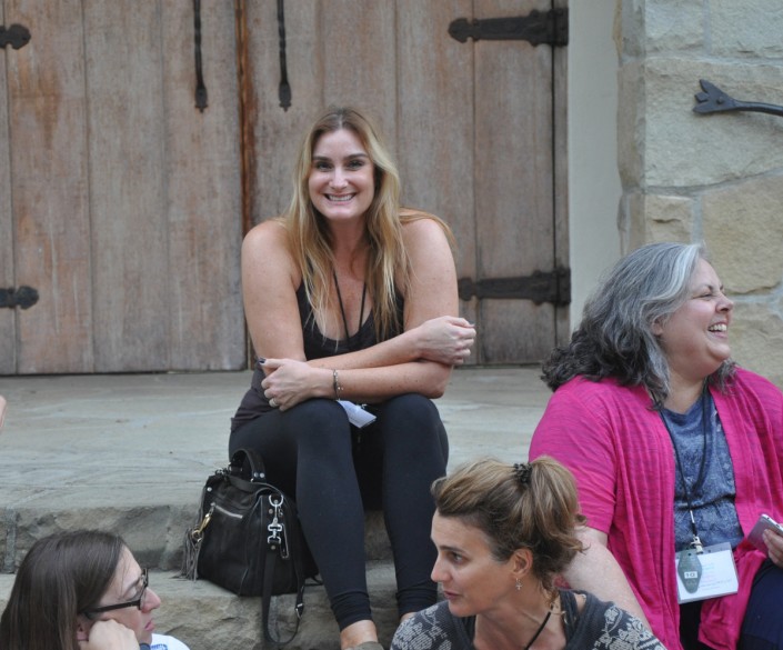 a woman wearing all black yoga clothing seated on steps and smiling at the camera while other women chat around her