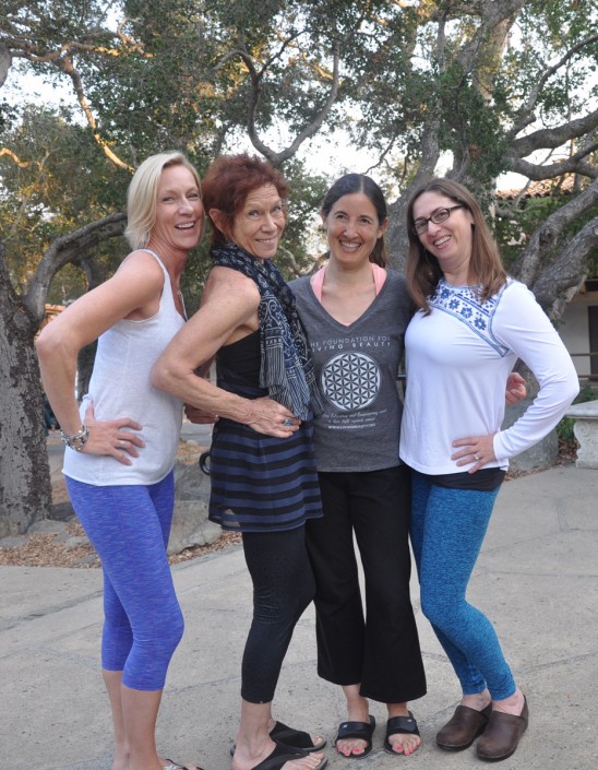 four women posing with their hands on their hips and smiling in front of large trees