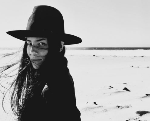 a woman in a black hat, black jacket with long black hair standing on a barren landscape