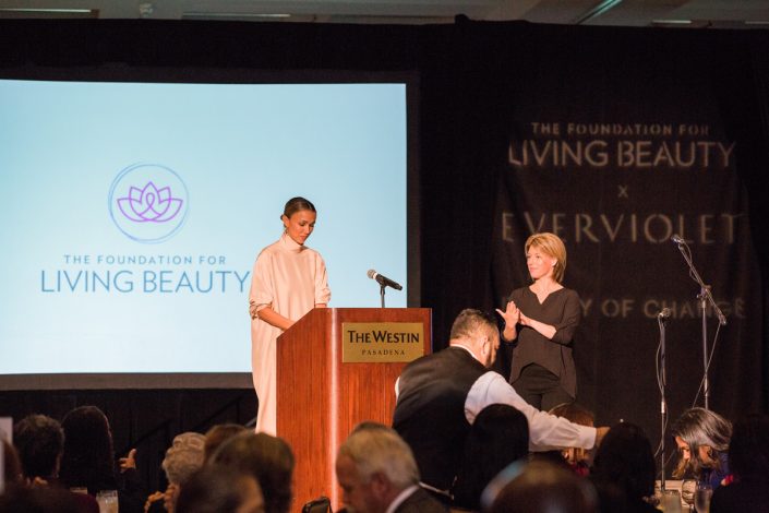 a woman in white standing at a podium in front of a projector screen showing the Foundation for Living Beauty logo