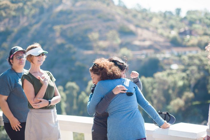 a woman in a blue sweater hugging a woman in a gray shirt with two women looking on