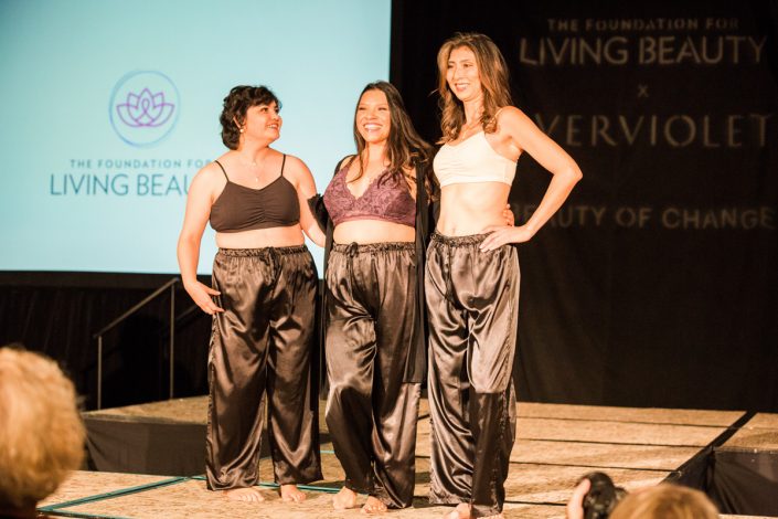 three women with their arms around each other standing on a runway in front of a screen with the Living Beauty logo on it