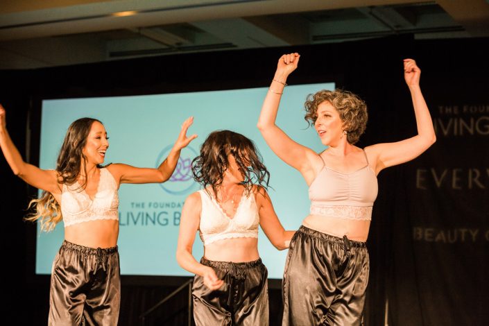 three women with their hands in the air standing in front of a projector screen showing the Living Beauty logo