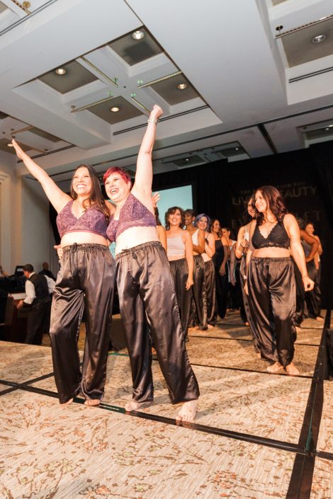 two women wearing purple bras and black drawstring pants standing on a runway with their hands in the air while other women line up behind them