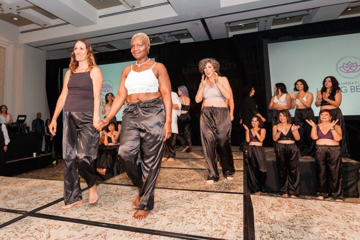 two women holding hands on a runway wearing similar outfits of tank tops and black drawstring pants while women behind them clap