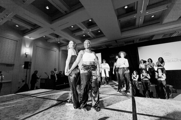 a black and white photo of two women on a runway standing back to back while other women behind them are clapping