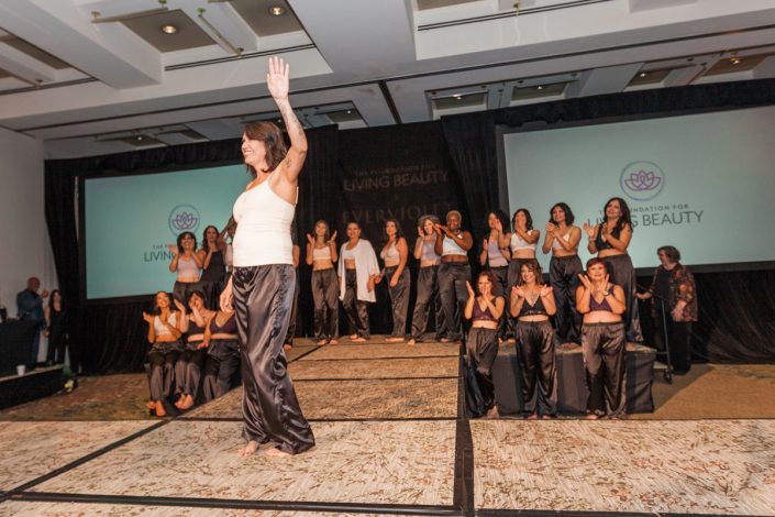 a woman wearing a white tank top and black pants standing on a runway waving while other women in similar outfits stand behind her and clap