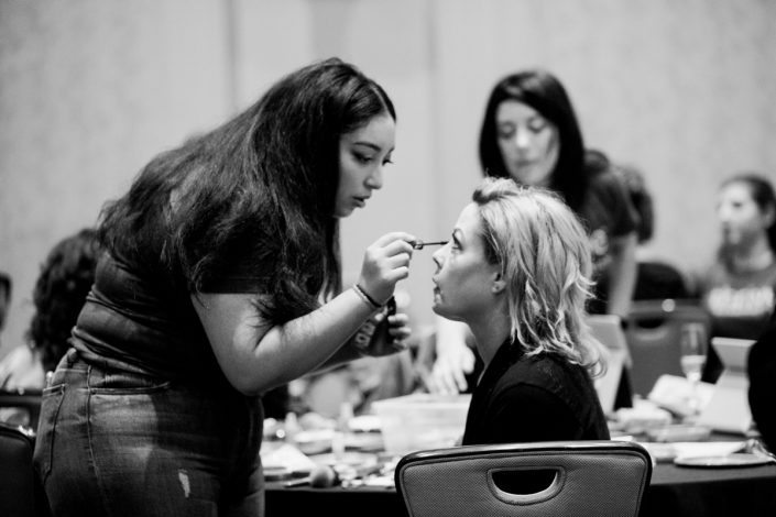 a black and white image of a woman standing applying eye makeup to a woman who is seated