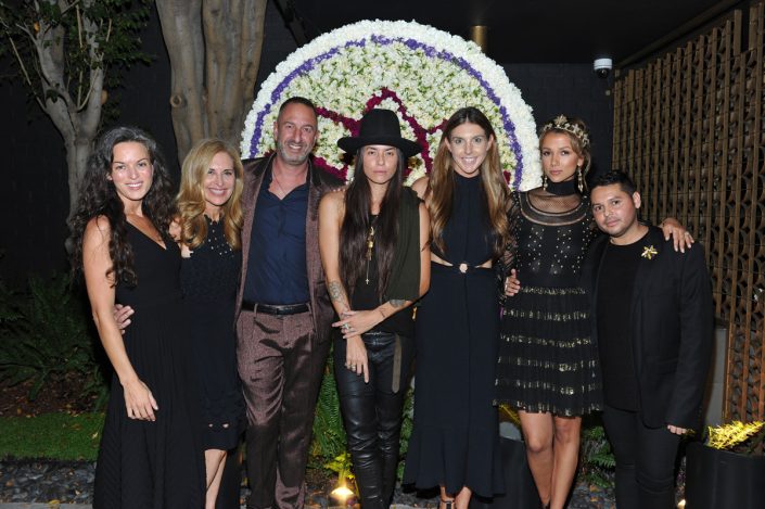a large group of men and women all dressed in black standing in front of a large floral arrangement of the Foundation for Living Beauty logo