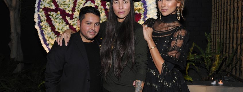 three people all dressed in black standing in front of a floral arrangement of the Foundation for Living Beauty logo