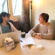 two women seated at a table while one is drinking coffee with a laptop, tissue, and clipboard in front of them