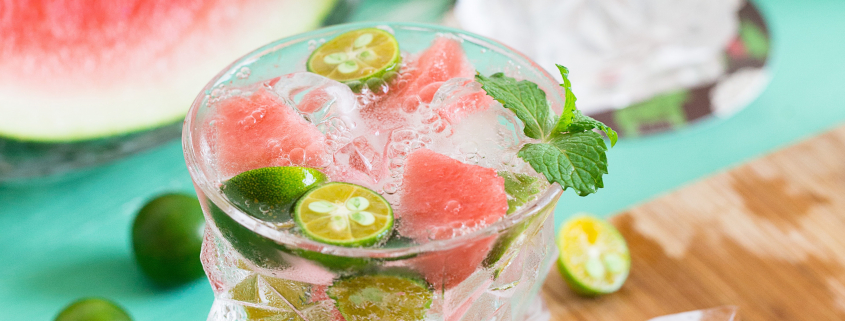 a glass filled with ice, mint and watermelon set on a cutting board with watermelon slices in the background