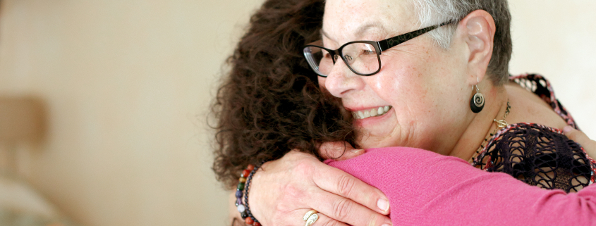 a woman with gray hair wearing glasses hugging a woman in a pink sweater whose face cannot be seen